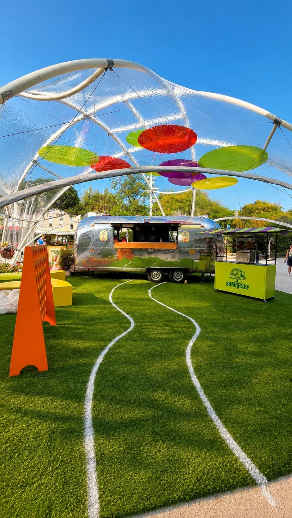 Caravane Airstream sous un dôme transparent, disques de couleur suspendus, comptoir vert de la marque sur la pelouse