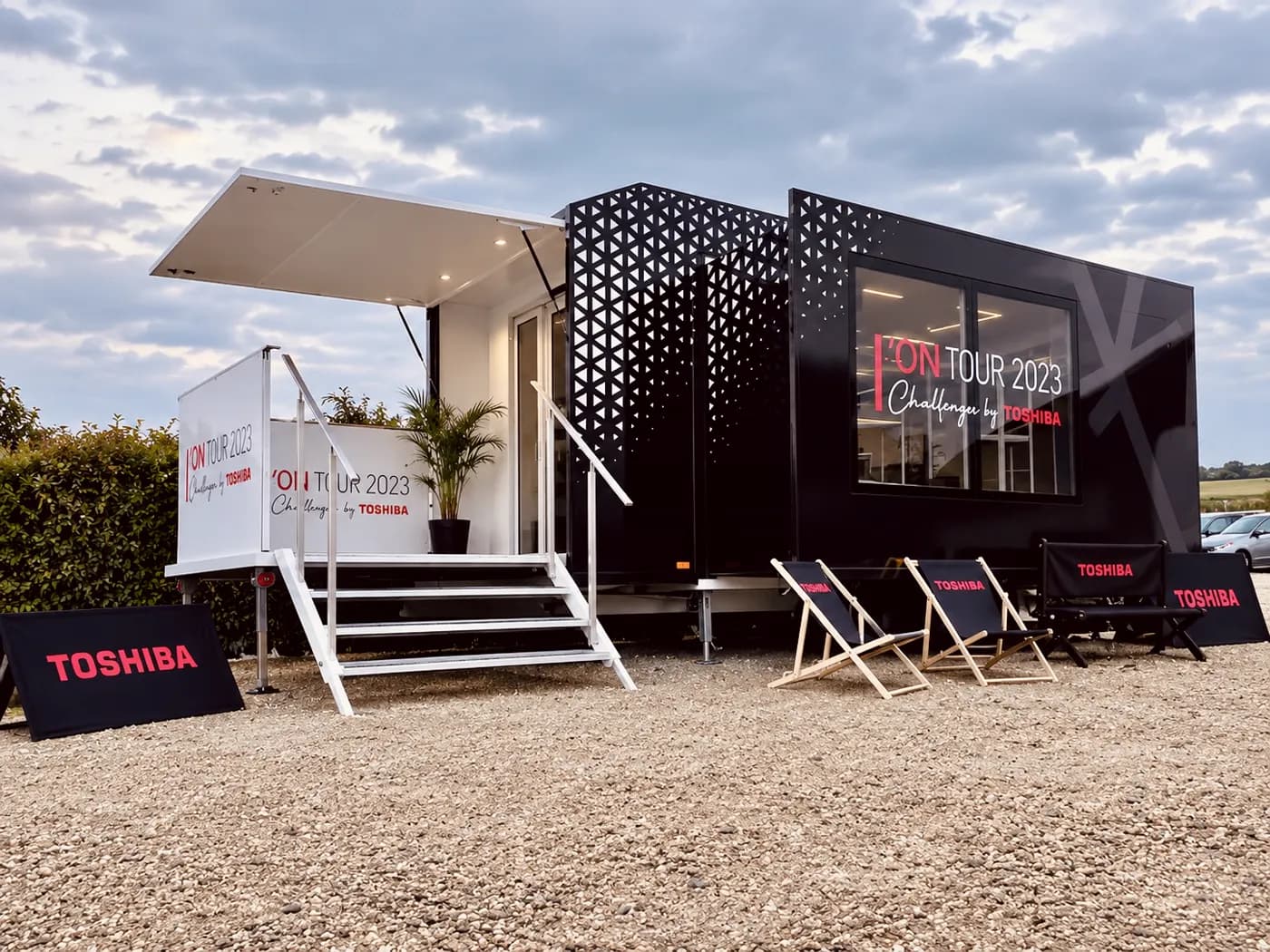 Showroom itinérant ouvert sur un parvis, façade perforée et mobilier d'accueil devant l'escalier