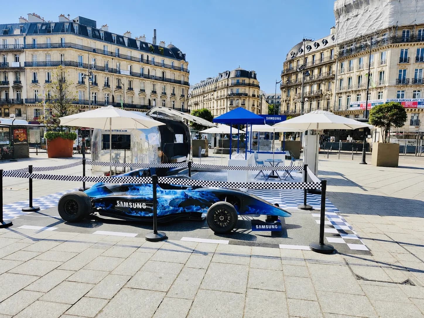 Monoplace de course habillée d'un décor bleu et caravane Airstream installées sur un sol à damier, sous des parasols, au milieu des passants sur une place bordée d'immeubles haussmanniens