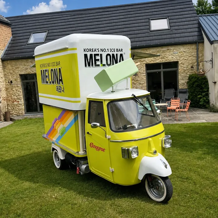 Triporteur jaune Melona Ice Bar sur une pelouse devant une ferme en pierre