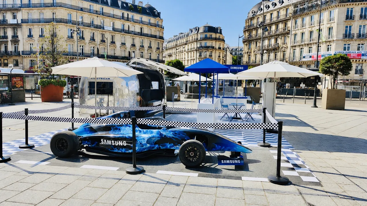 Monoplace de Formule 1 aux couleurs Samsung posée sur un damier bleu et blanc, sur une place bordée d'immeubles haussmanniens